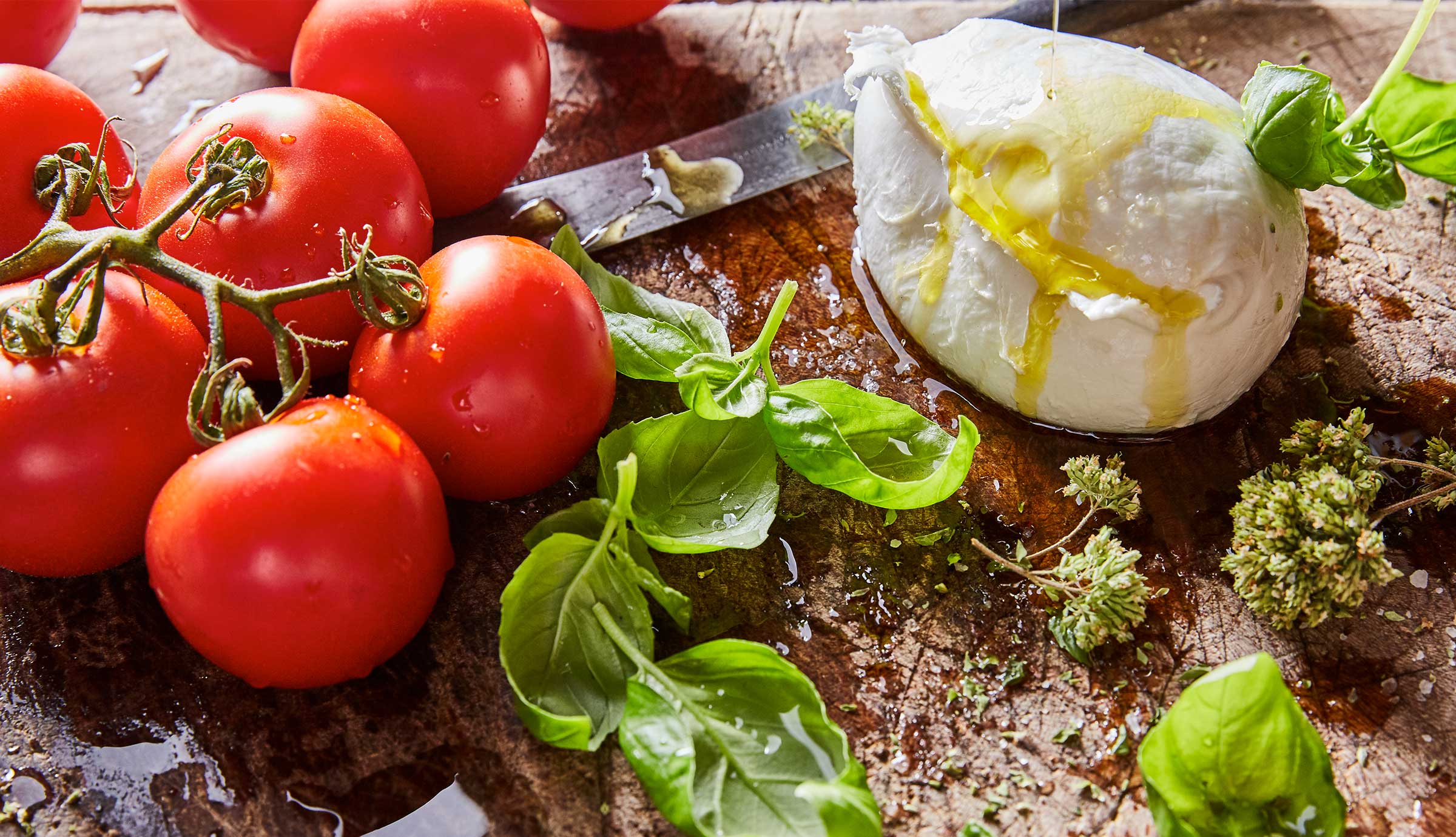 Tomatoes, mozzarella cheese, basil and oregano sit on a wood surface